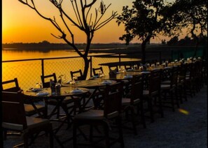 Un área de comedor al aire libre con mesas y sillas, con vista a un cuerpo de agua durante la puesta de sol.