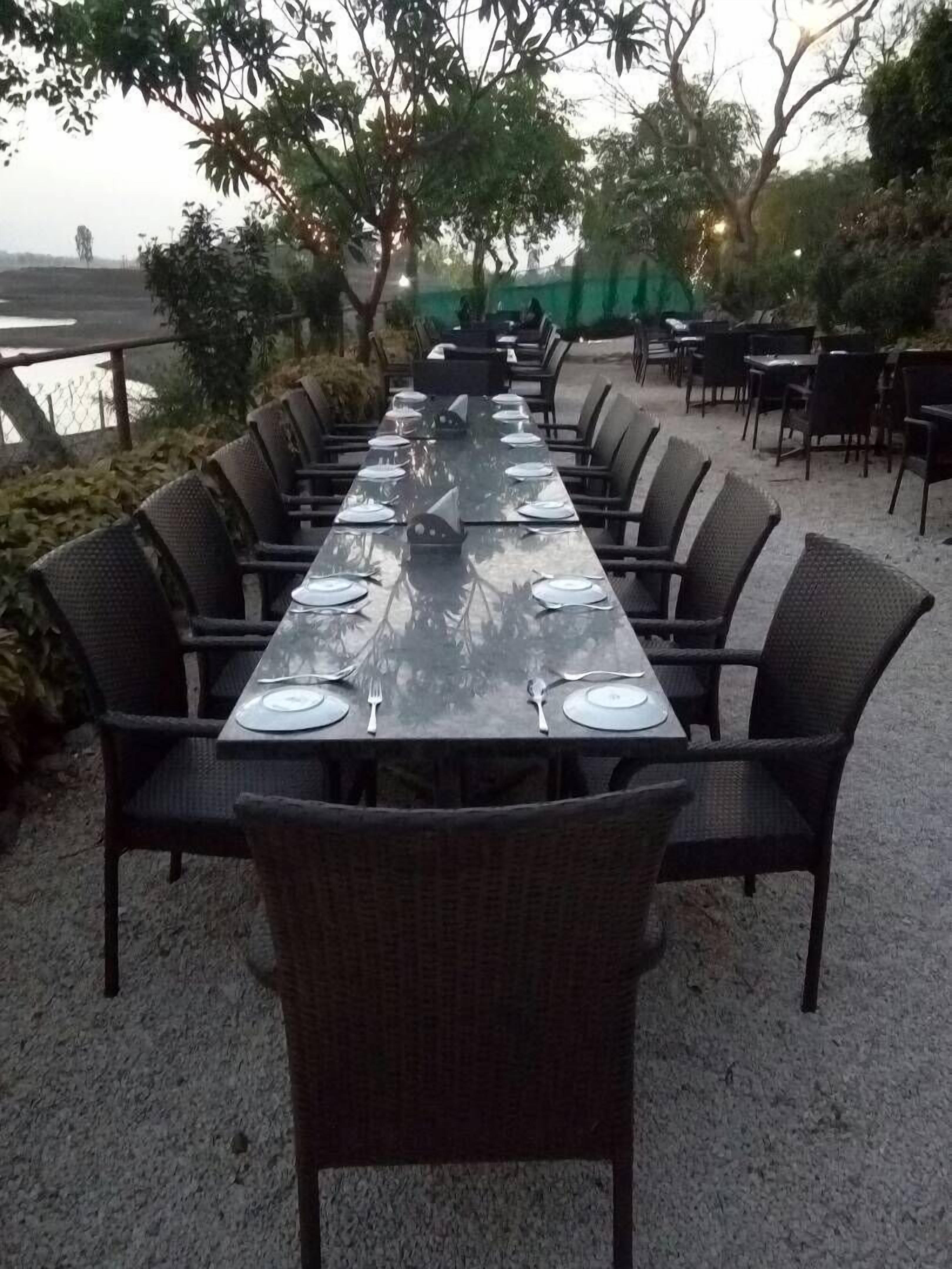 A long dining table set outdoors with chairs, surrounded by greenery and a view of a body of water.