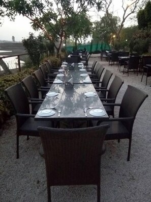 Una larga mesa de comedor al aire libre con sillas, rodeada de follaje y vistas a un cuerpo de agua.