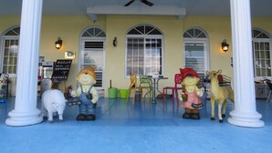Un espace extérieur abrité, doté d’un ventilateur de plafond, de colonnes blanches et d’un sol bleu. On y trouve deux tables avec des chaises, une petite statue représentant une personne avec un chien, et un panneau avec du texte.