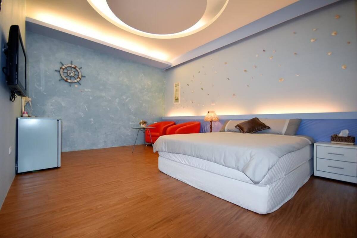 Une chambre moderne avec un grand lit, un petit bureau, une chaise rouge et un téléviseur placé sur le mur de gauche.