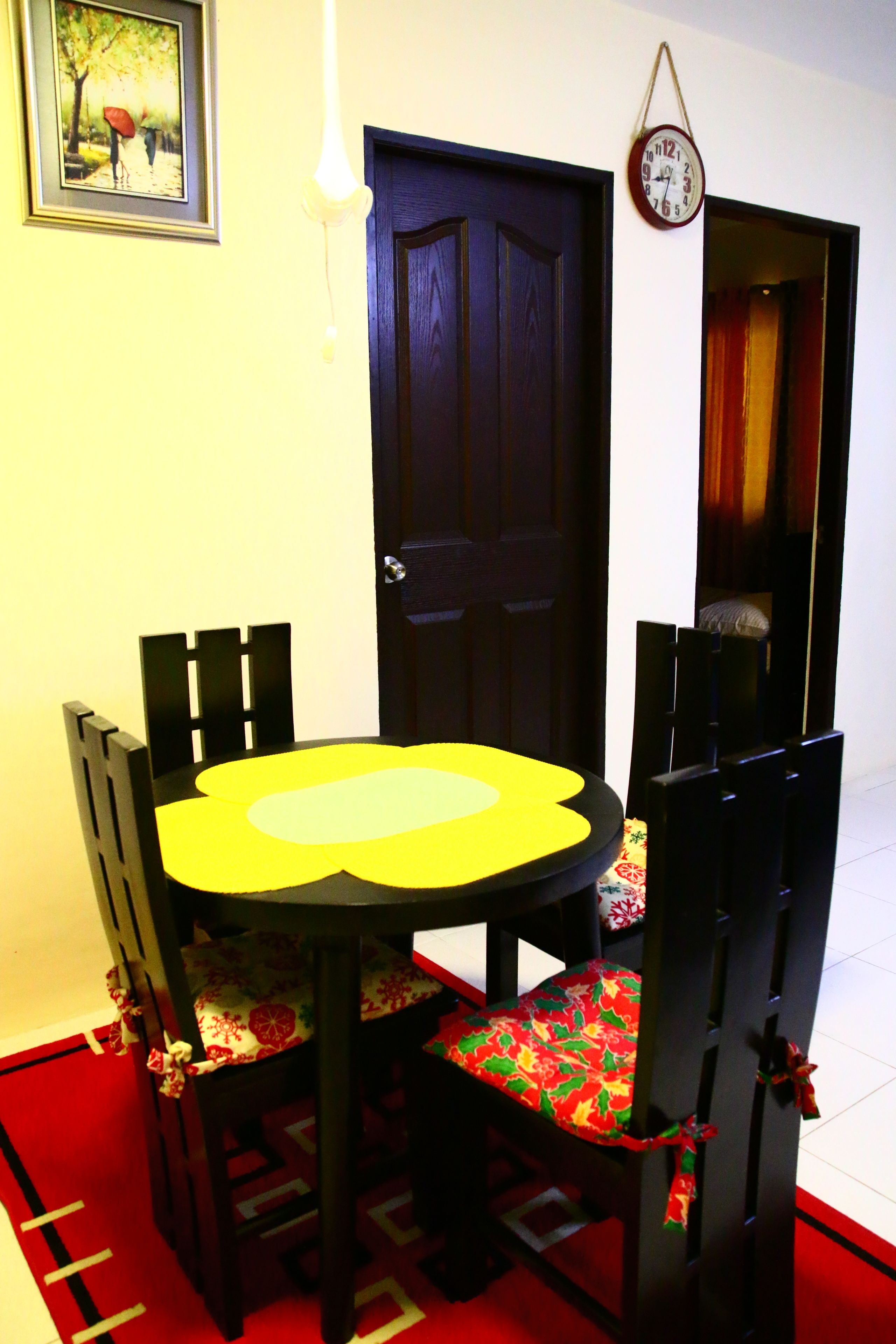 Un comedor con una mesa redonda y cuatro sillas, una puerta, un reloj de pared y un cuadro enmarcado.