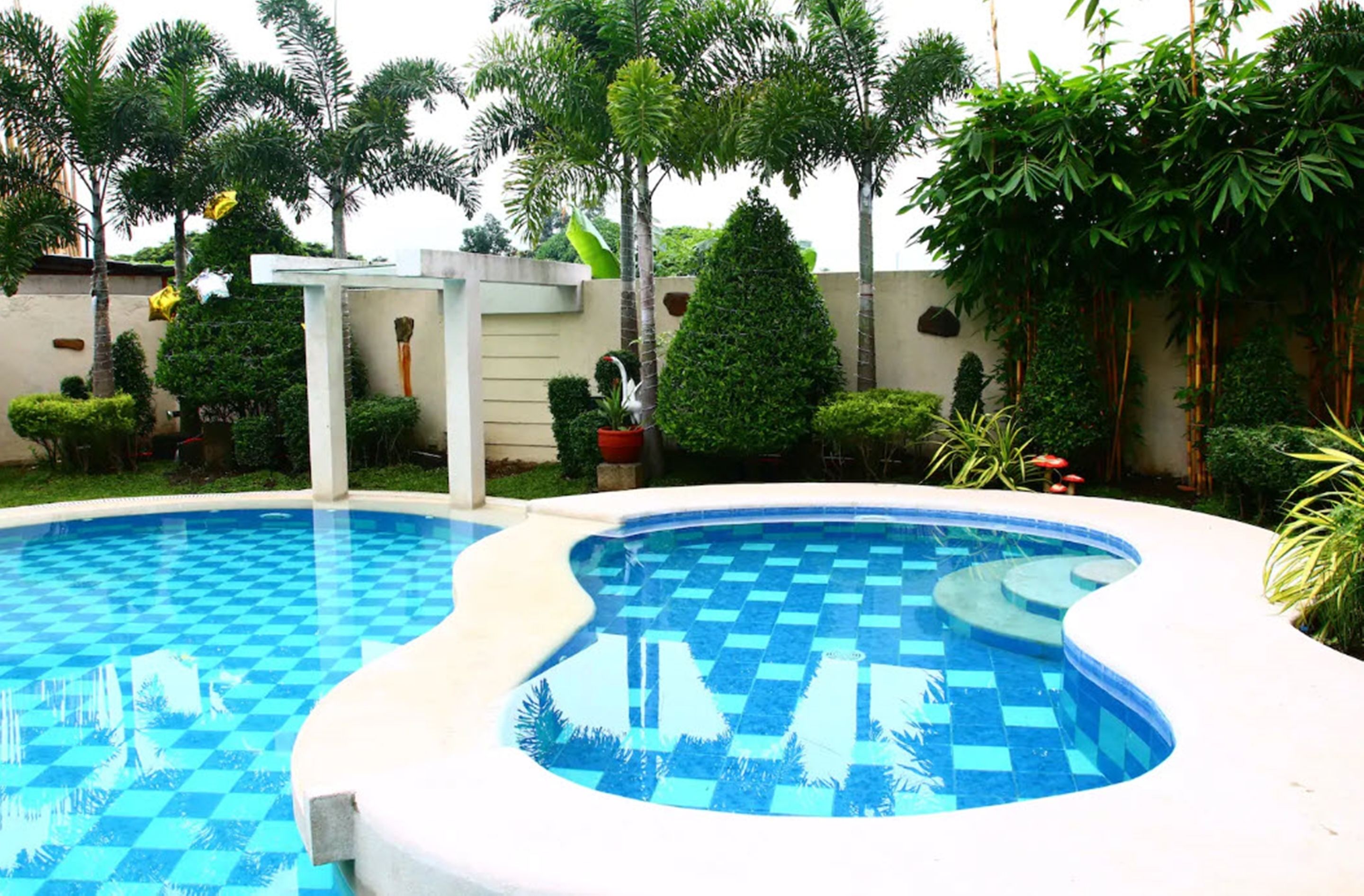 A swimming pool with a checkered pattern, surrounded by various plants and a stone wall.