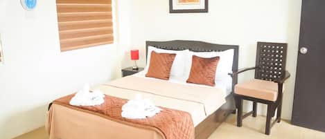 A neatly made bed with a brown bedspread and two pillows, a wooden chair, a red lamp, and a framed picture on the wall.