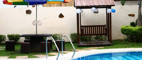 Una piscina con diseño a cuadros, una pérgola de madera con luces esféricas blancas y un paraguas colorido.