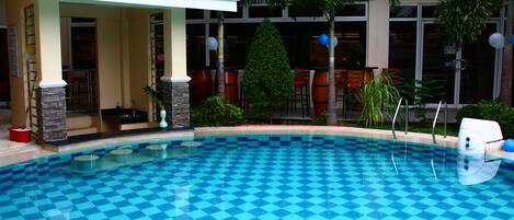 Una piscina con diseño a cuadros, un área de estar cubierta y un restaurante al fondo.