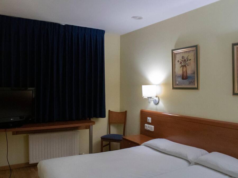 A hotel room with a bed, a chair, a desk with a television, and a wall-mounted lamp.