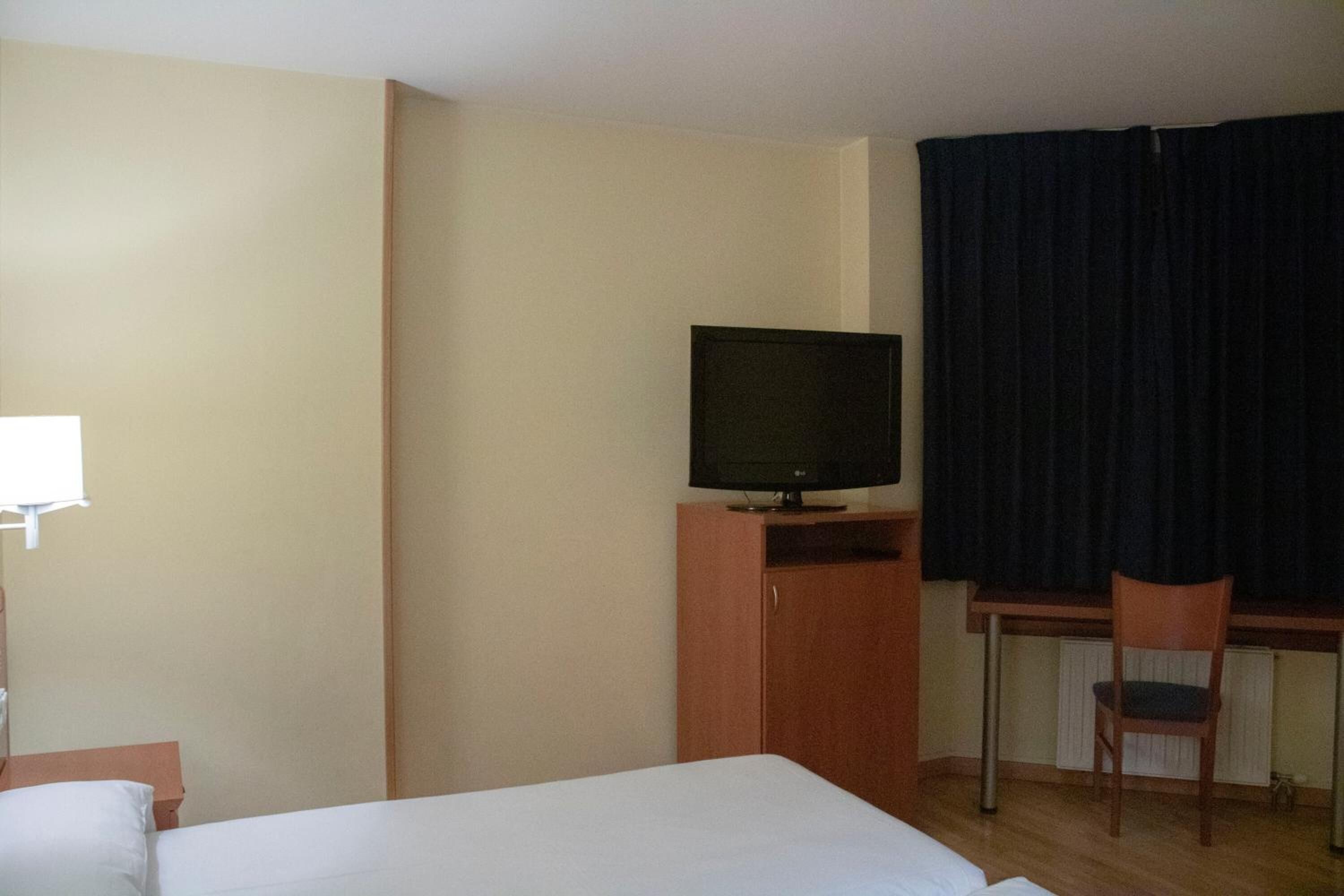 Ein Hotelzimmer mit Bett, Fernseher auf einer Holzkommode, Schreibtisch mit Stuhl und einem Fenster mit Vorhängen.