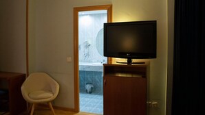 Een hotelkamer met een televisie op een houten standaard, een witte stoel en een badkamer die door een geopende deur zichtbaar is.