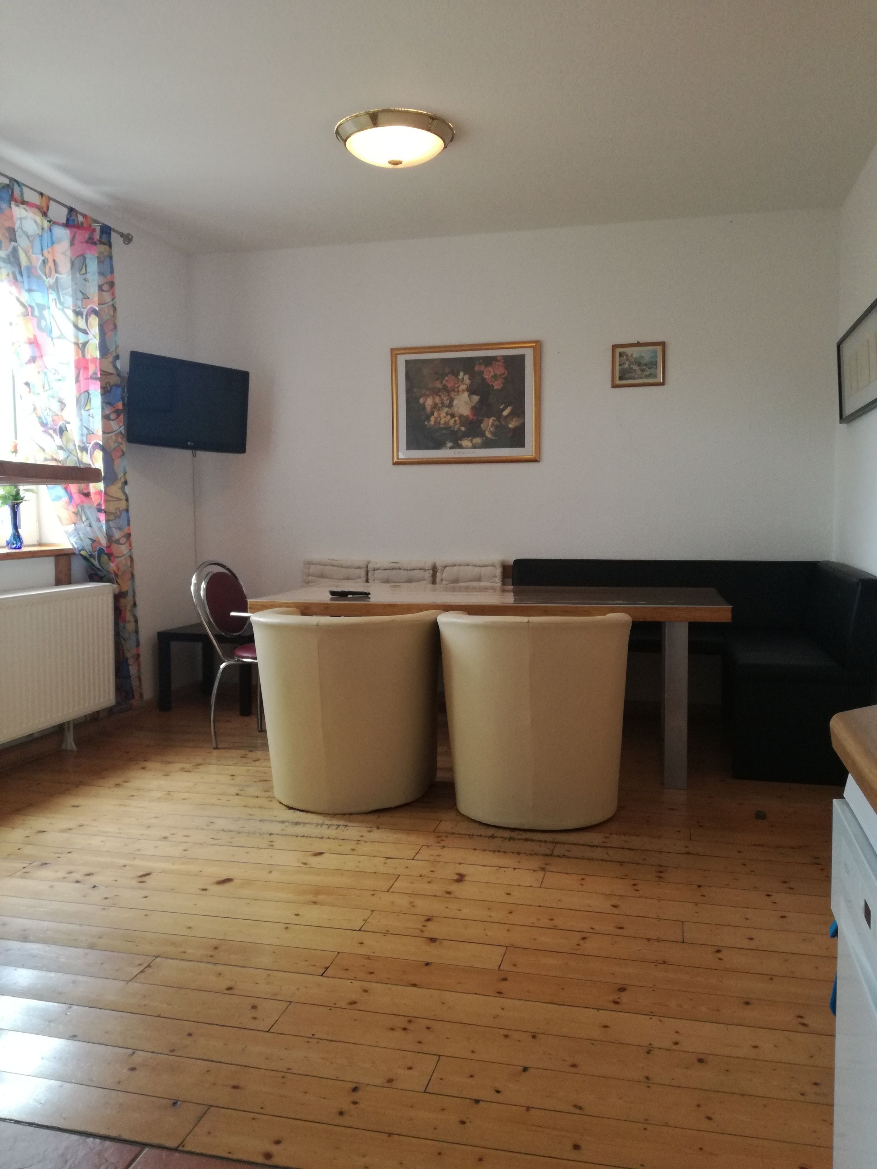 A room with a wooden floor, a table with two stools, a black sofa, a TV mounted on the wall, and two framed pictures.