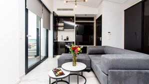 A modern living room with a grey sofa, a round coffee table, and a small side table. There is a large window on the left side, and a kitchen area is visible in the background. - Crystal villas & suites (Kassandra)
