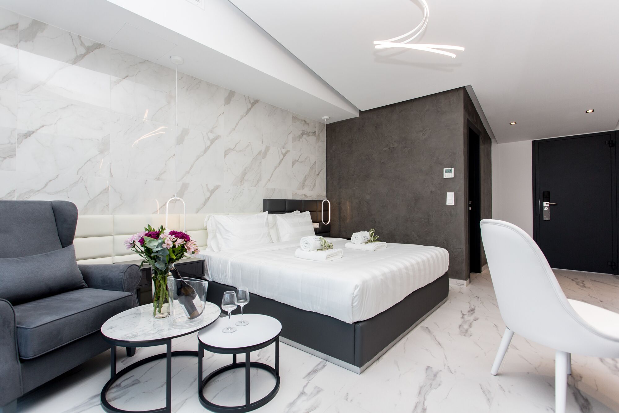 Un dormitorio moderno con cama, sofá gris, dos mesitas y una silla blanca.