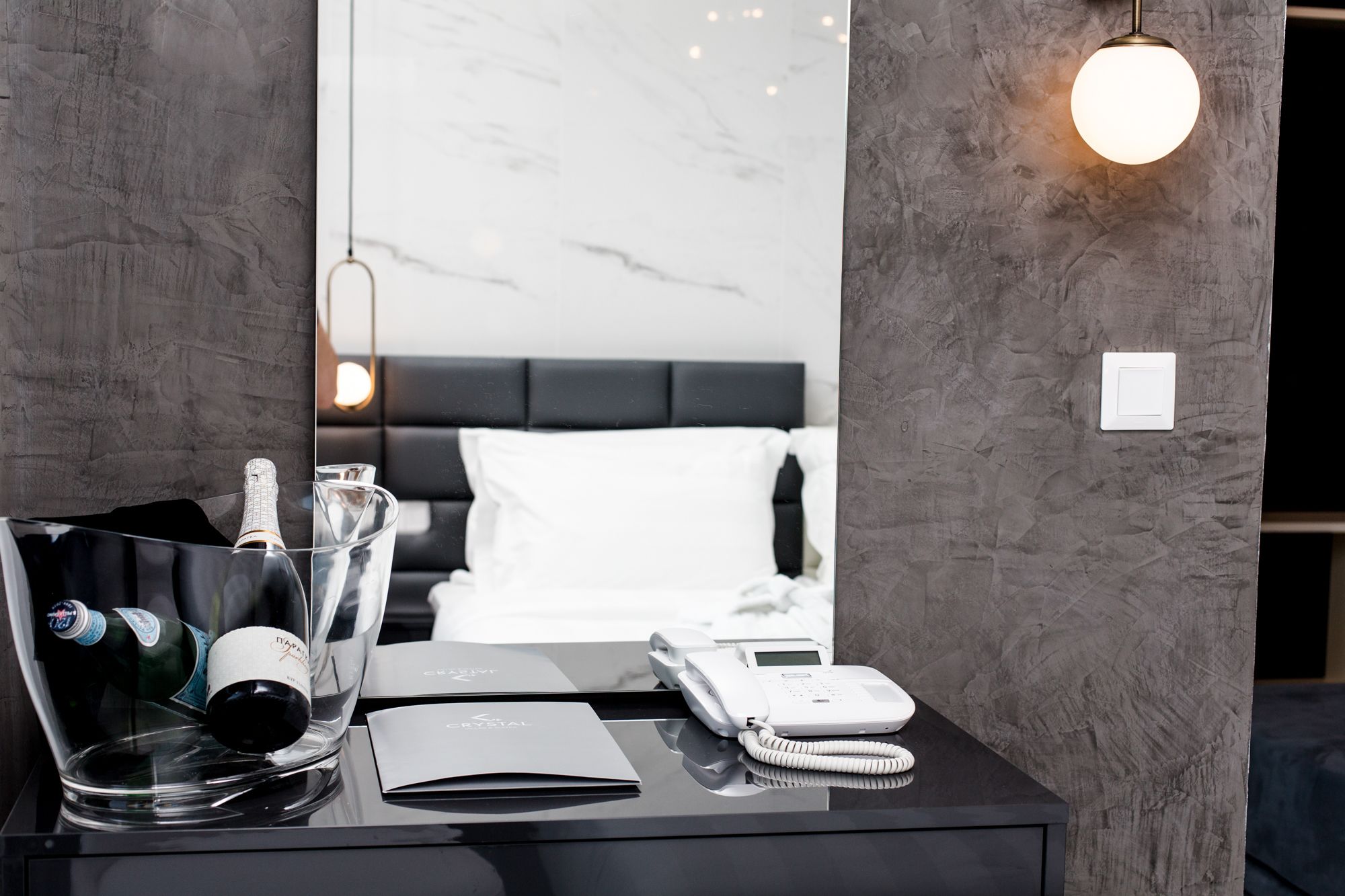 A hotel room with a black bedside table, a bed with white bedding, a modern telephone, and a mirror reflecting the room.