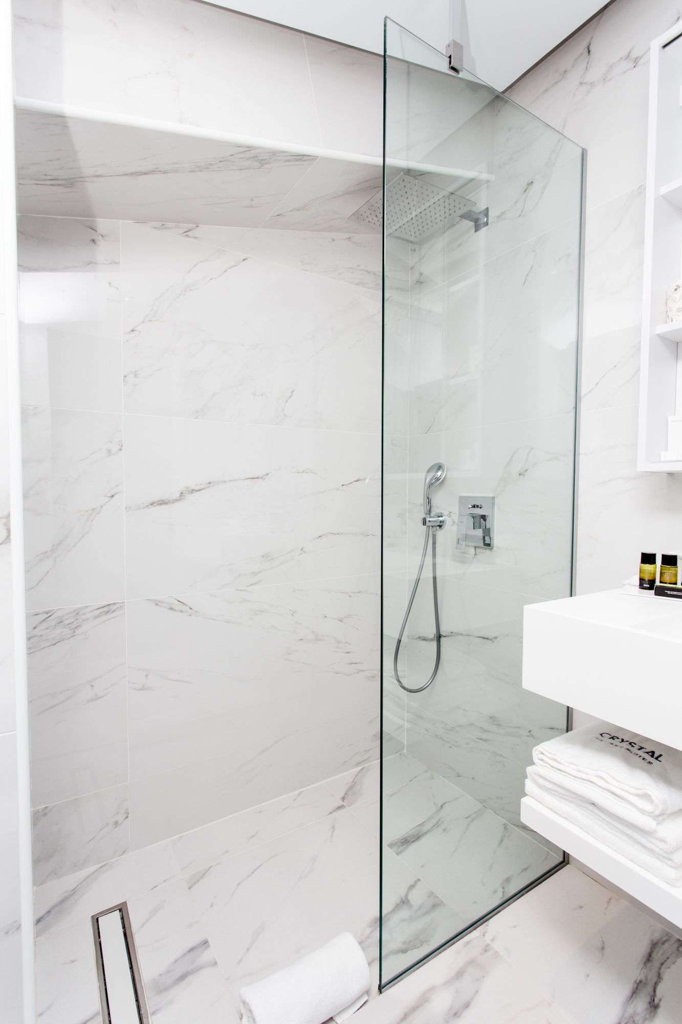 Une salle de bain moderne avec une douche à parois de verre, des carreaux de marbre, un lavabo blanc et des serviettes soigneusement pliées.
