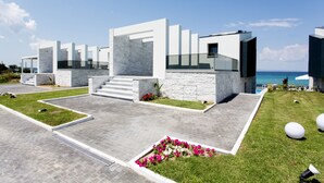 A modern building with a stone facade, a set of stairs leading to a covered entrance, and a view of the ocean. - Crystal villas & suites (Kassandra)