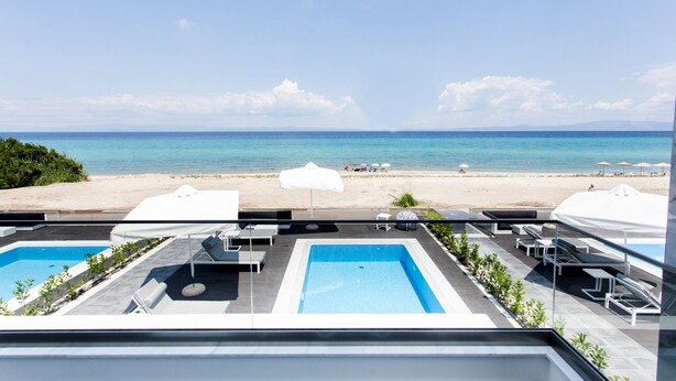 A rooftop pool area with two pools, lounge chairs, and umbrellas, overlooking a beach with umbrellas and people. - Crystal villas & suites (Kassandra)