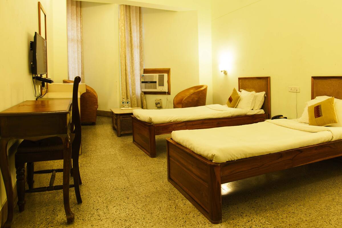 Une chambre d’hôtel avec deux lits en bois, un bureau, une chaise, un climatiseur et un téléviseur.