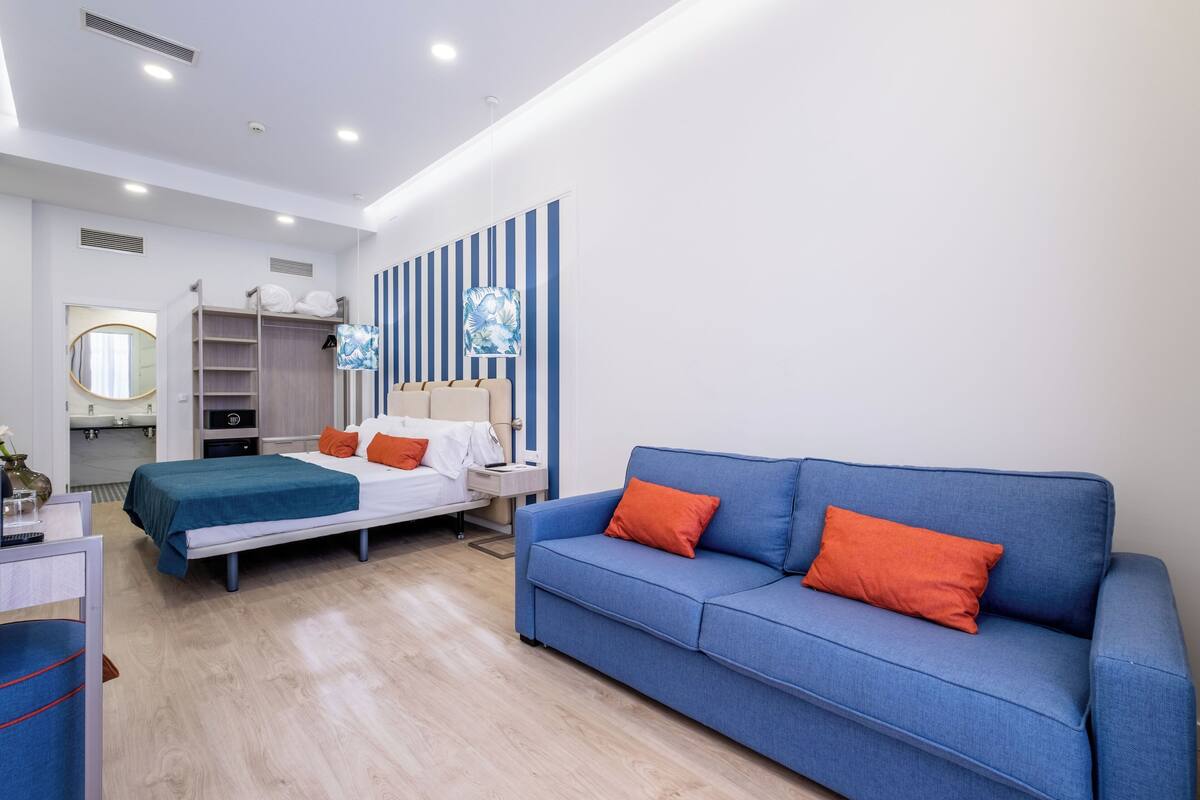 A modern hotel room with a bed, a blue sofa, a vanity with a mirror, and a striped wall.