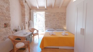 A cozy room with a bed, a wicker chair, a small table with a pitcher, and a view of the outside. - Historic Piazza House & Romantic Garden (Hvar)