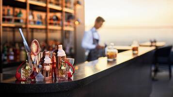 A bar area with various bottles and glasses on the counter, shelves with bottles in the background, and a person in the distance.