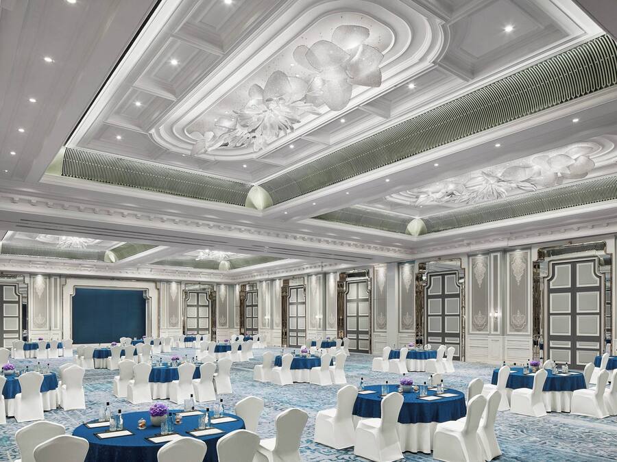 A large, elegantly decorated banquet hall with round tables and white chairs, featuring intricate ceiling designs and large windows.