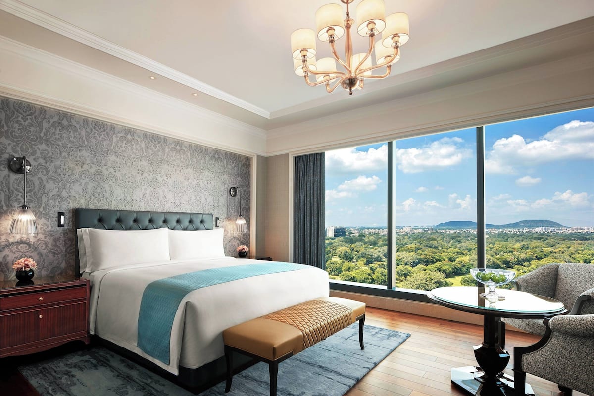 A modern hotel room with a large bed, a view of a cityscape, and a balcony with a table and chair.