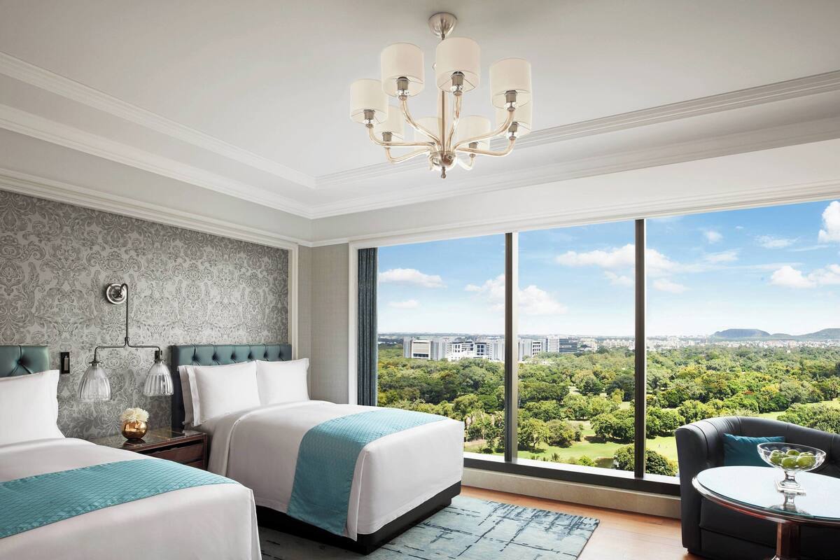 A hotel room with two beds, a chandelier, a large window with a city view, and a small table with a bowl of fruit.