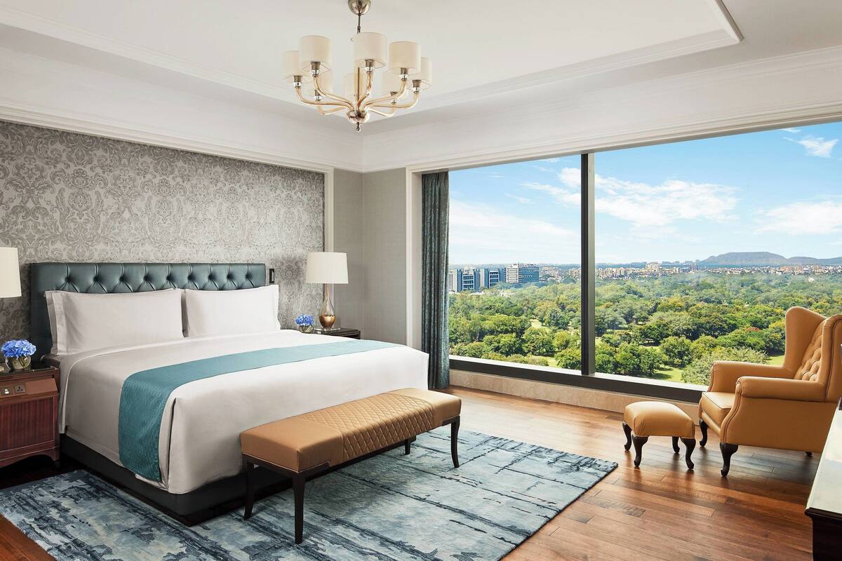 A modern hotel room with a large bed, a view of a cityscape, a chandelier, and a city park view.