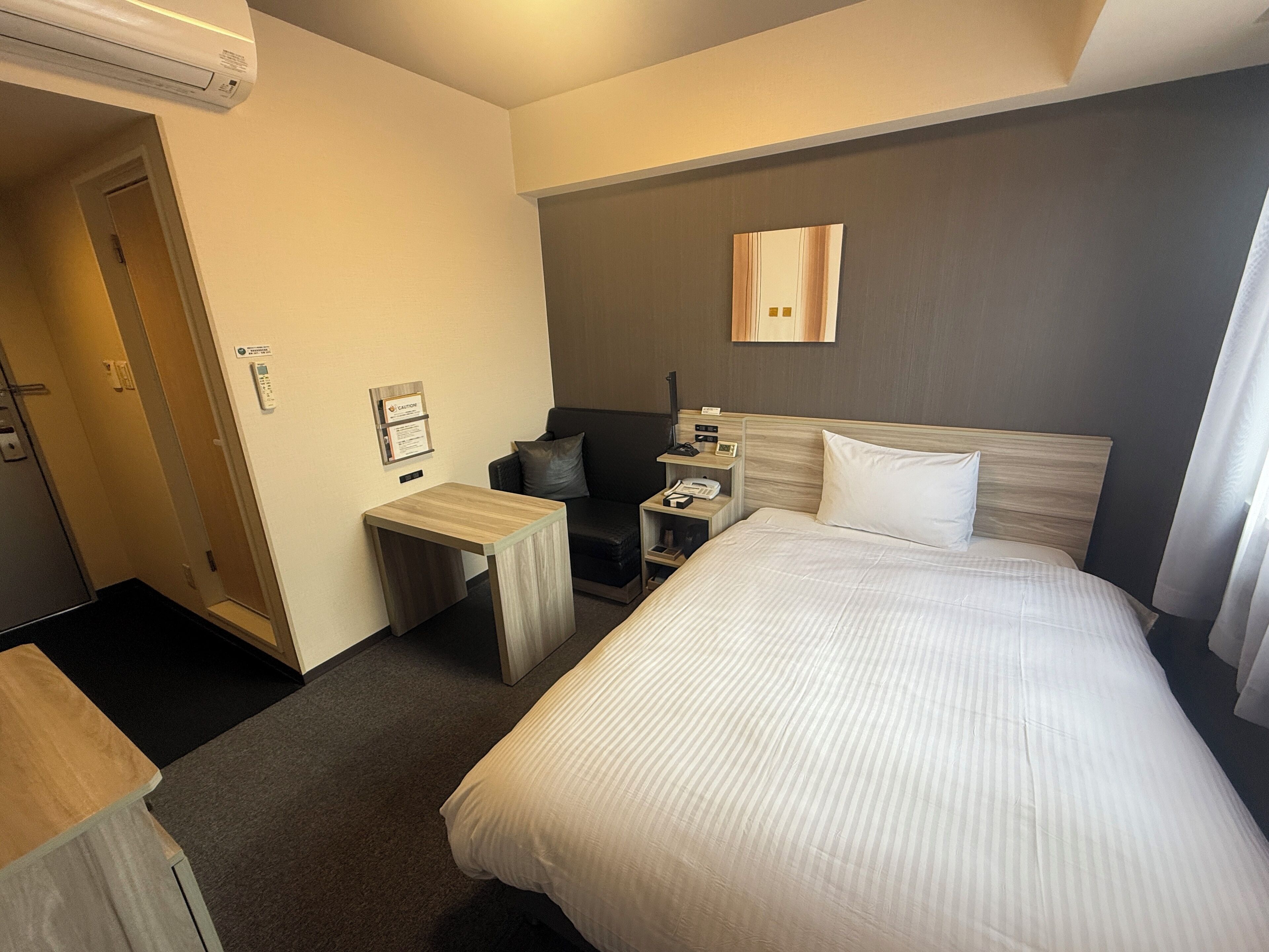 A hotel room with a bed, bedside table, nightstand, and a small seating area with a chair and a sofa.