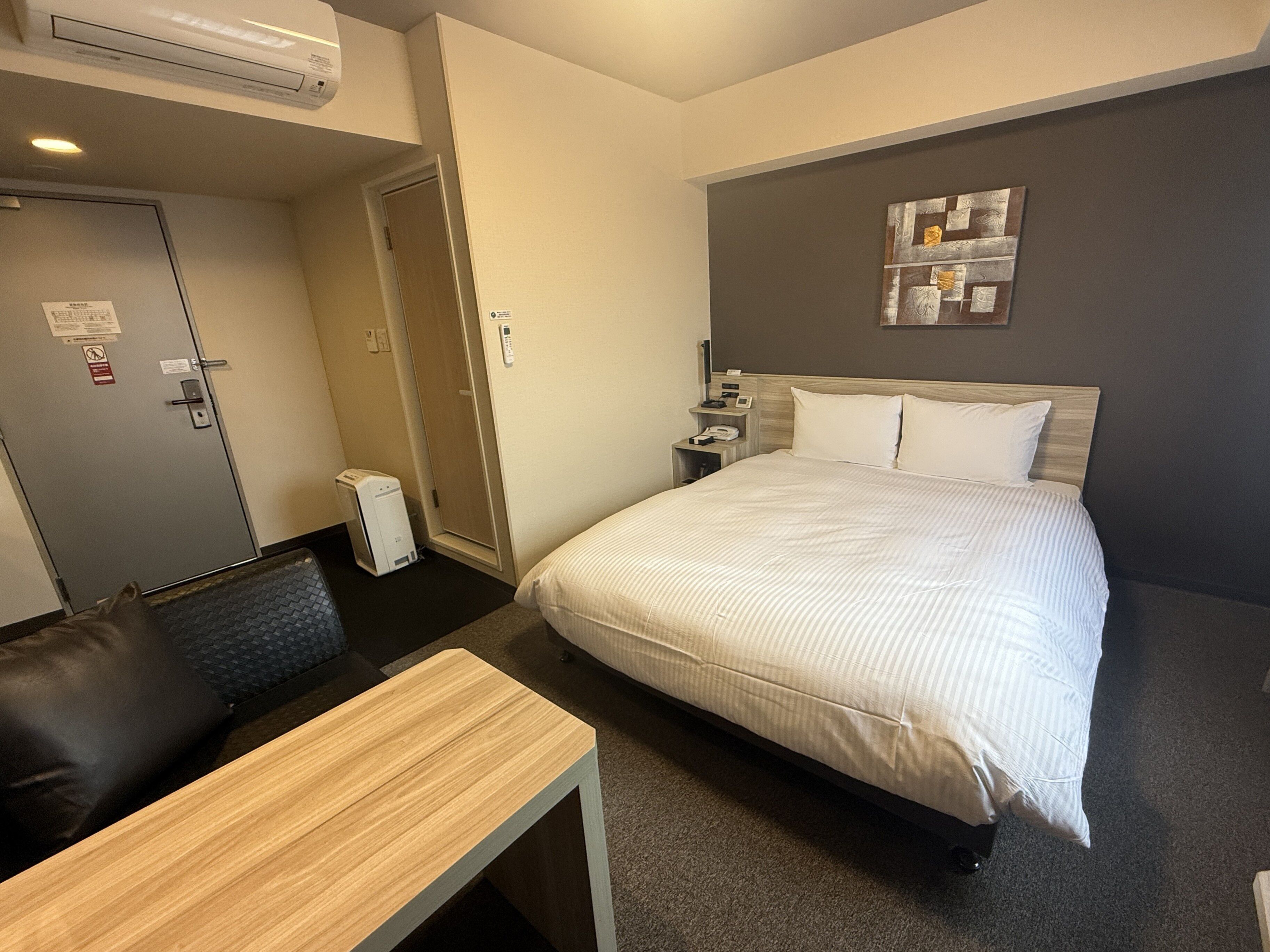 A hotel room with a bed, a desk, a chair, a nightstand, and a wall-mounted artwork.