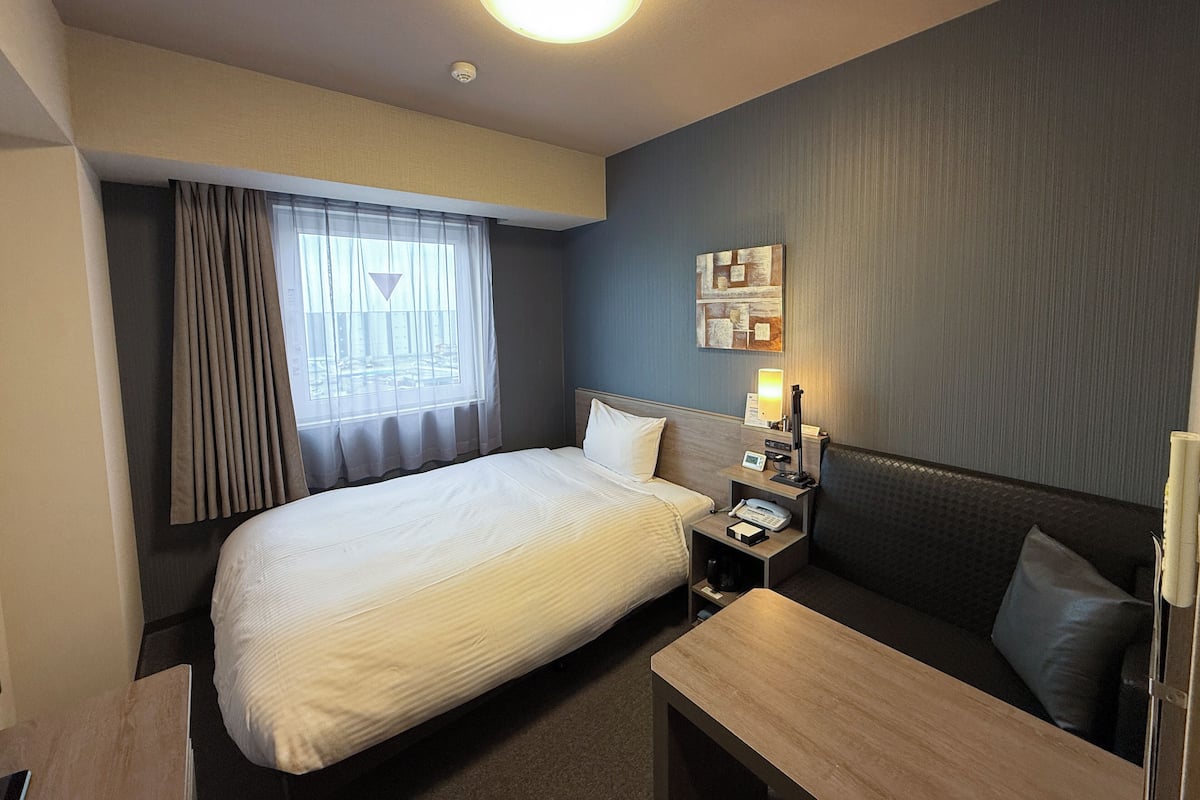 Une chambre d’hôtel avec un lit, une table de chevet avec une lampe, un petit bureau, un canapé et une fenêtre avec des rideaux.