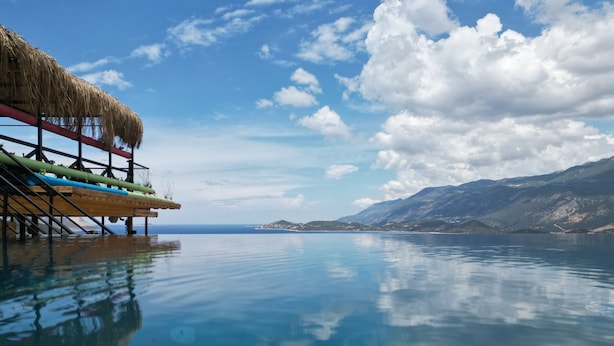 A tranquil infinity pool with a thatched-roof structure and kayaks on the left, overlooking a calm sea and distant mountains. - Stone Hill Kas (Kas)