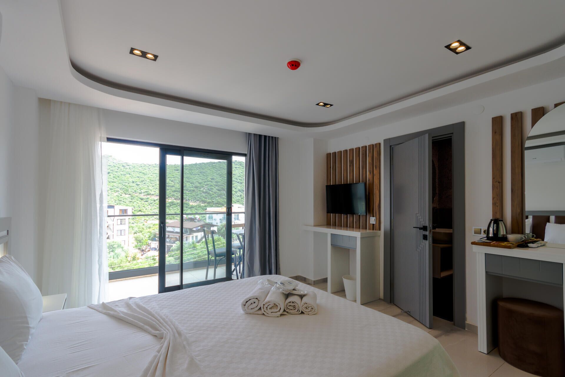 Une chambre d’hôtel comprenant un lit, un téléviseur fixé sur un panneau en bois, un bureau avec une chaise et un balcon offrant une vue sur la verdure.