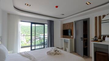 Une chambre d’hôtel comprenant un lit, un téléviseur fixé sur un panneau en bois, un bureau avec une chaise et un balcon offrant une vue sur la verdure.