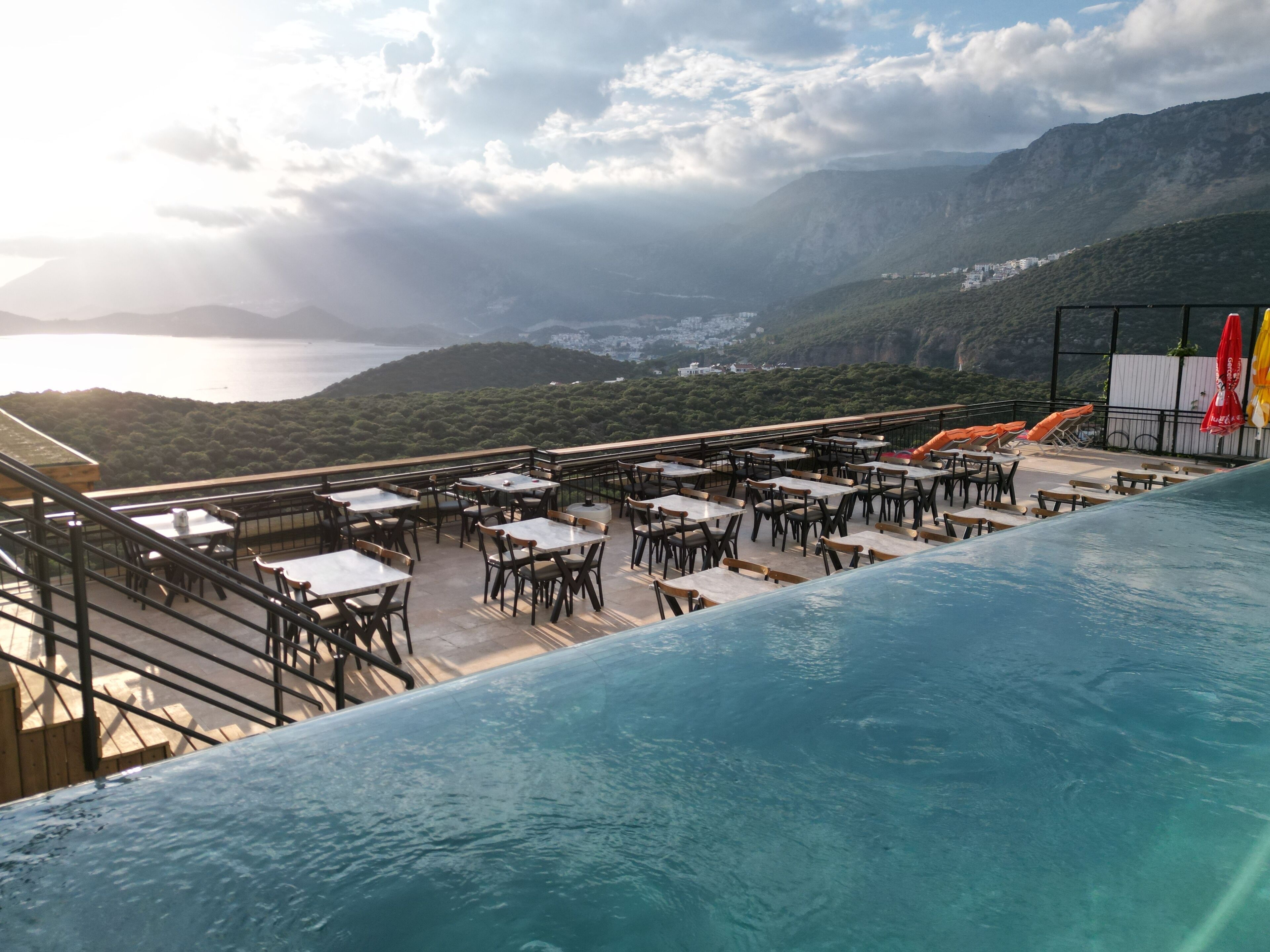 A rooftop pool area with tables and chairs, overlooking a mountainous landscape and a body of water.