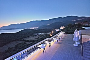 Terraço com mesas e cadeiras com vista para a paisagem costeira e montanhas ao fundo.