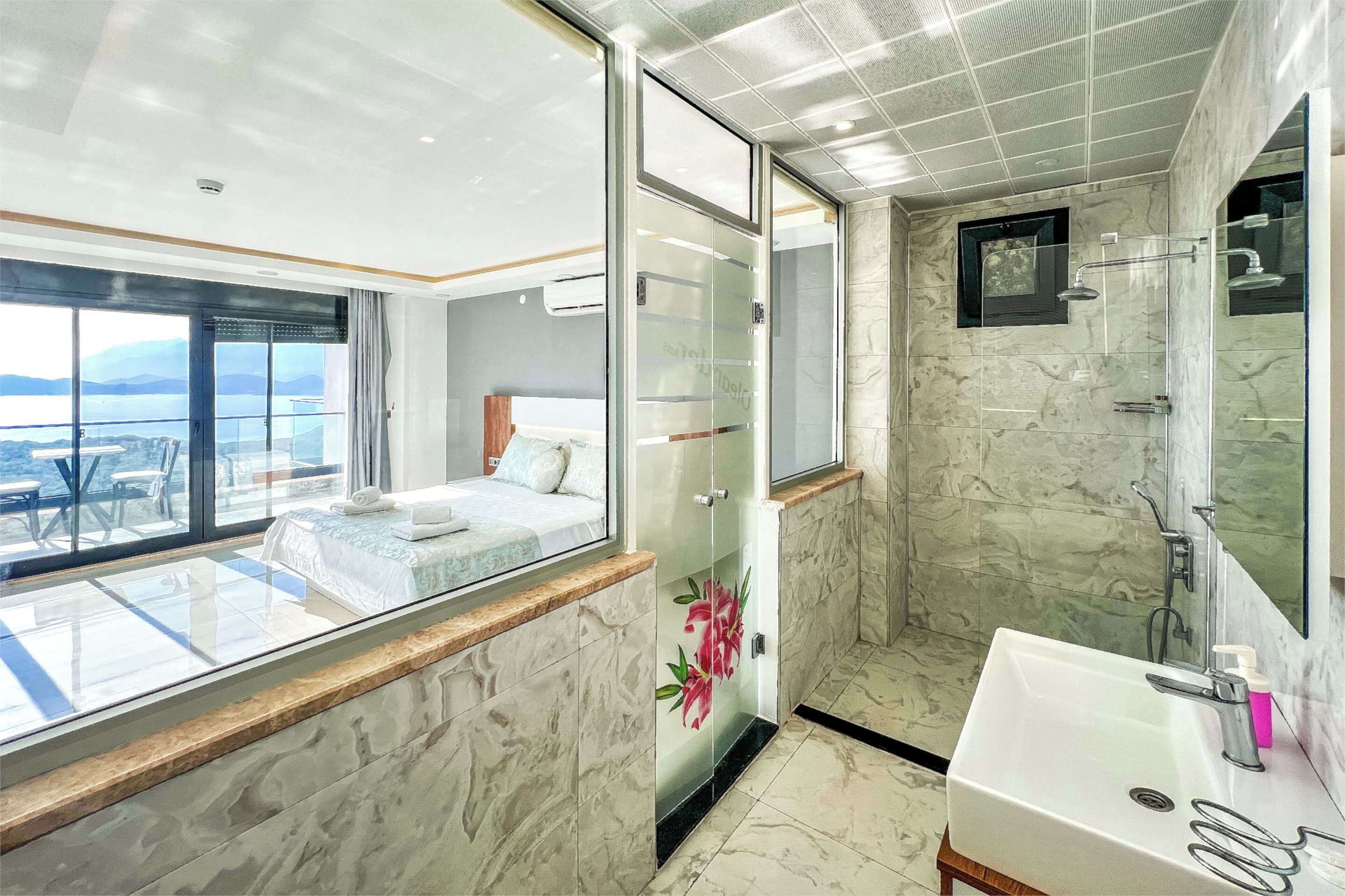 A modern hotel room with a balcony, a large bed, a glass shower enclosure, and a bathroom with a marble sink and mirror.