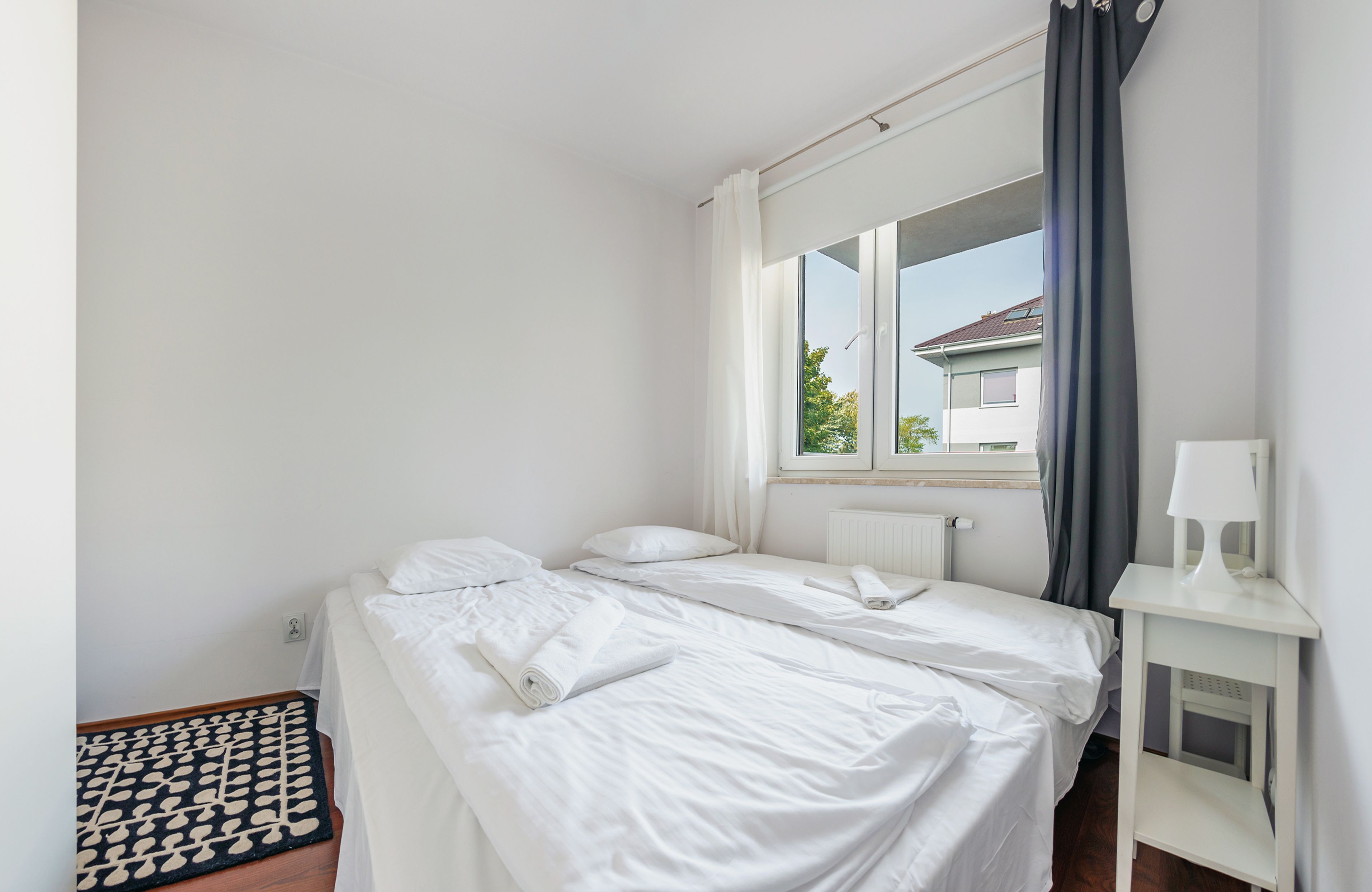A single bed with white linens, a small bedside table with a lamp, and a window with curtains.
