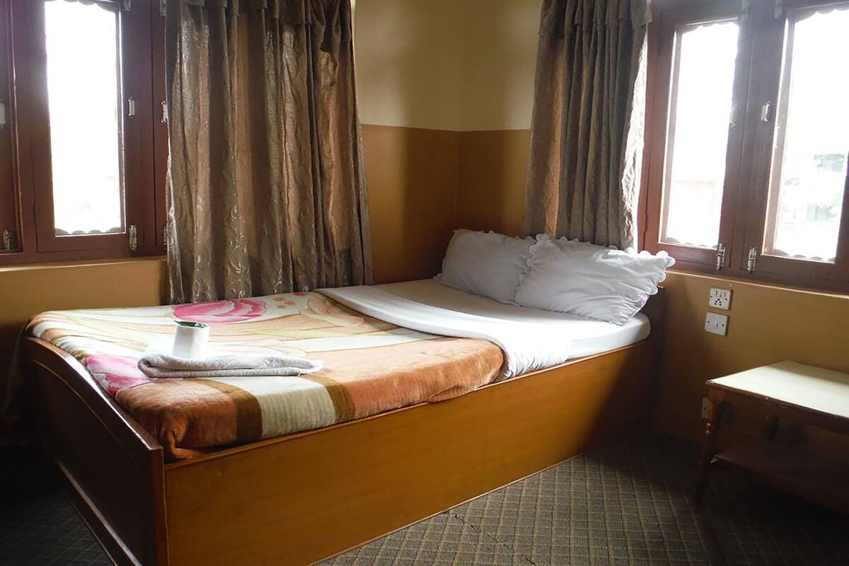 A single bed with a wooden frame, a white mattress, and a patterned bedspread. There is a small bedside table with a white surface and a drawer. The room has two windows with brown curtains, and the walls are painted in a light color.