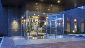 Uma entrada de hotel moderna com fachada de vidro, uma placa com a inscrição "HOTEL ROUTINN" e um interior bem iluminado visível pelas portas.