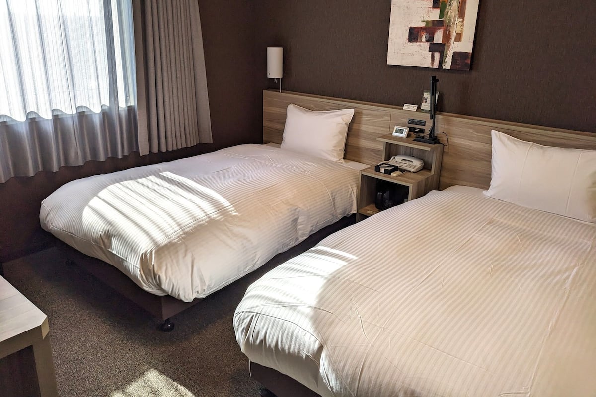A hotel room with two beds, a nightstand with a phone and lamp, a painting on the wall, and a window with curtains.