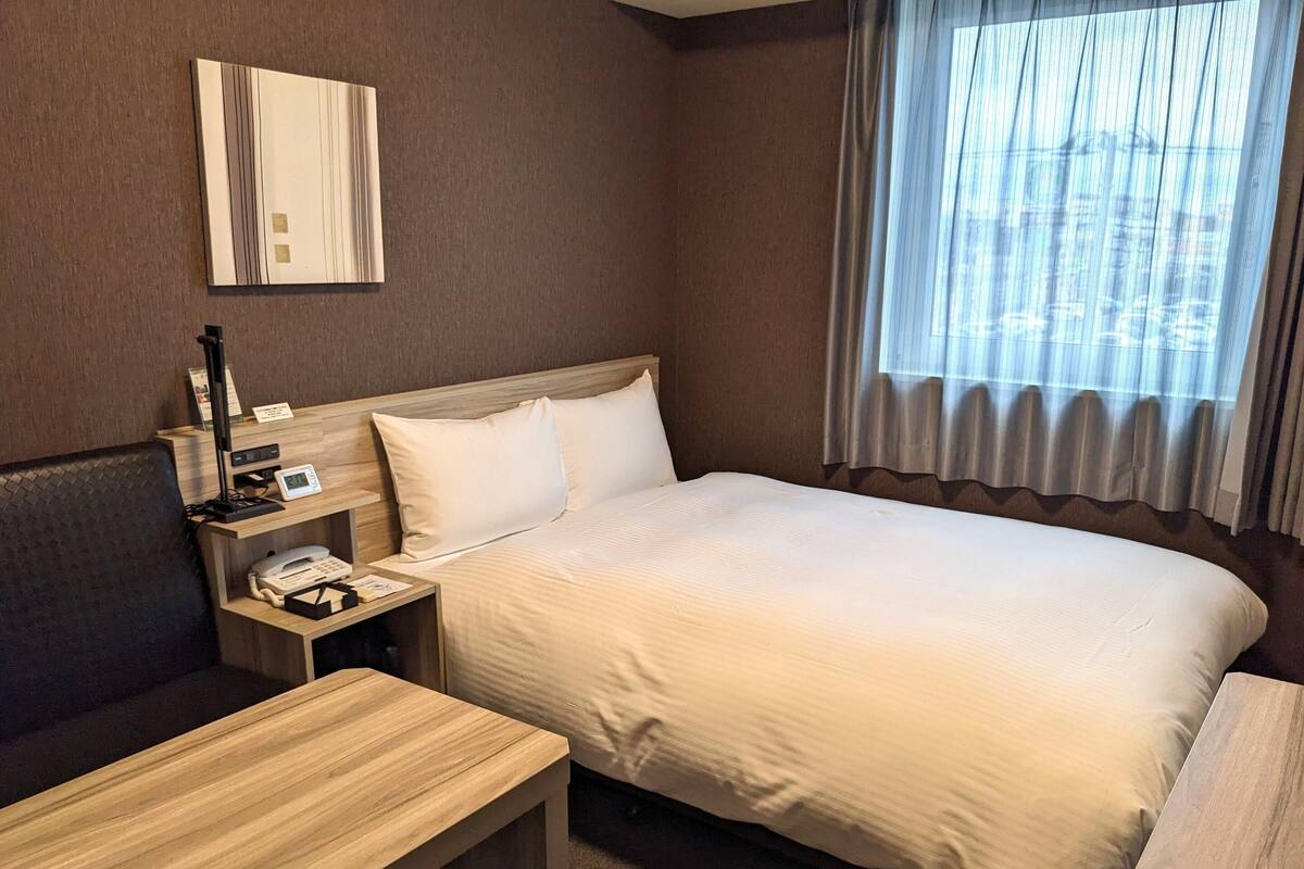 A hotel room with a bed, a desk, a chair, a bedside table, a phone, and a window with curtains.