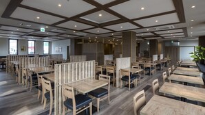 Um restaurante moderno com mesas e cadeiras de madeira, tetos altos com vigas aparentes e grandes janelas que permitem a entrada de luz natural.