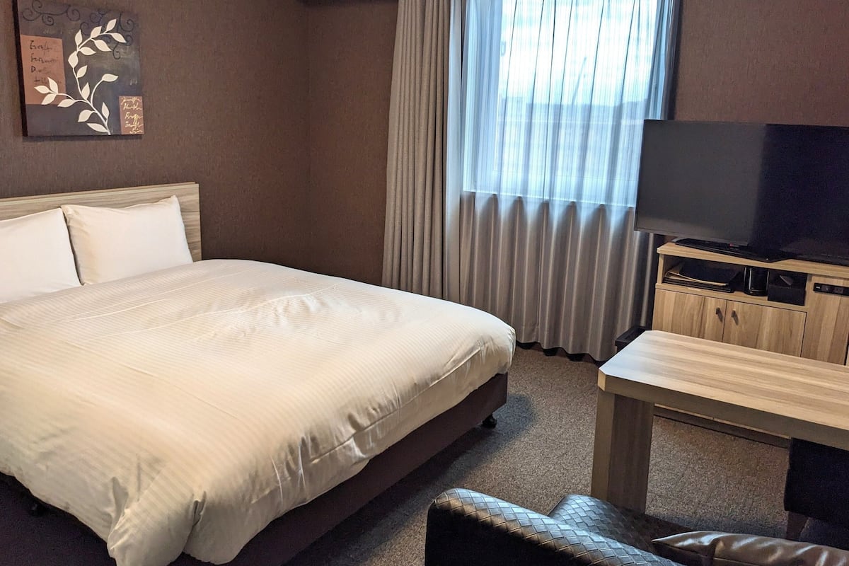 A hotel room with a large bed, a television on a wooden stand, a small table, and a window with curtains.