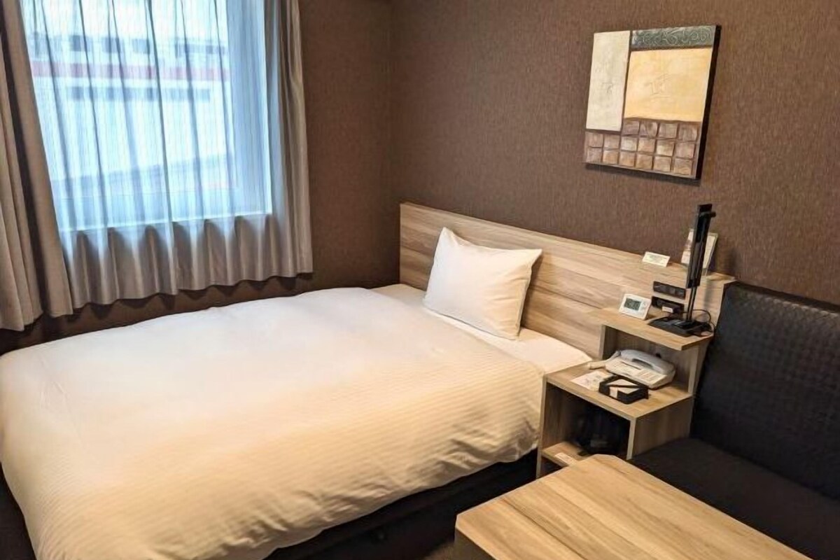 Una habitación de hotel con cama, mesita de noche con lámpara, pequeño escritorio y sofá.
