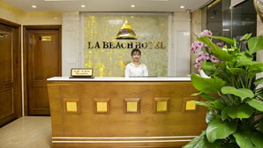 Taulell de recepció a l'Hotel La Beach, amb un recepcionista, un senyal de recepció i una planta gran en test.
