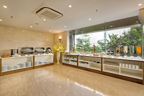 A modern buffet setup with multiple serving stations, a large window offering a view of greenery, and a clean, polished floor. - La Beach Hotel (Da Nang)