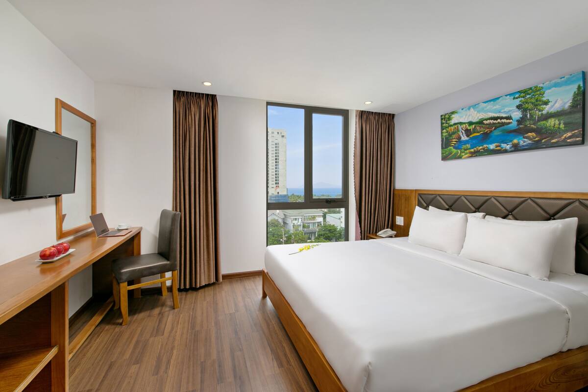 A hotel room with a bed, a desk with a laptop, a chair, a TV, and a window with curtains.