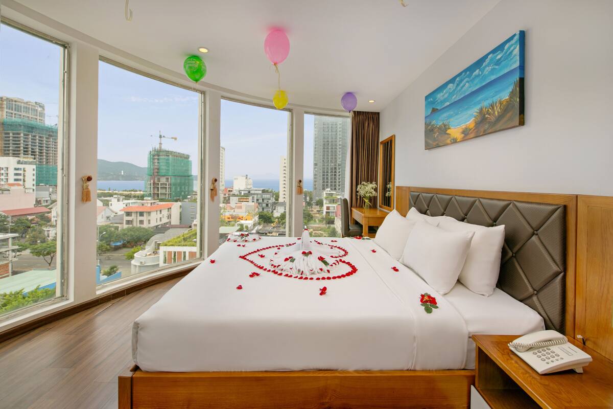 A hotel room with a large bed, a view of the city, and a painting on the wall.