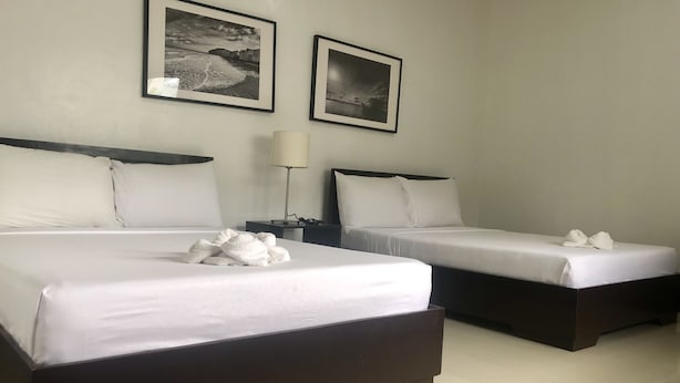 Quarto de hotel com duas camas, cada uma com roupa de cama e almofadas brancas, uma mesinha de cabeceira com abajur e dois quadros na parede.